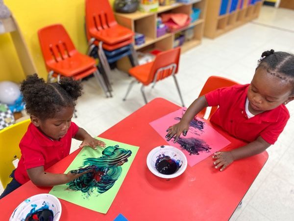 Two children painting at a red table in a classroom, with bowls of dark paint and colorful paper.