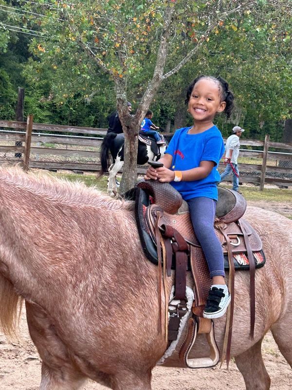 Child smiling on a brown horse at a ranch, wearing a blue shirt and helmet.