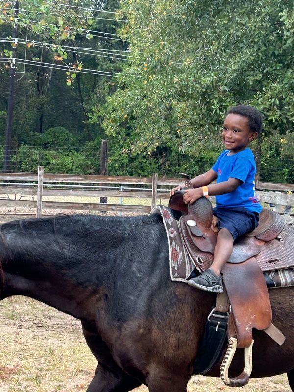 Child riding a dark horse outdoors on a ranch, smiling in a blue shirt.