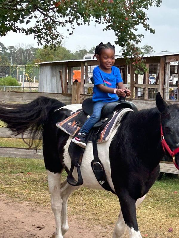 Child riding a black-and-white pony outdoors, wearing a blue shirt and smiling under trees