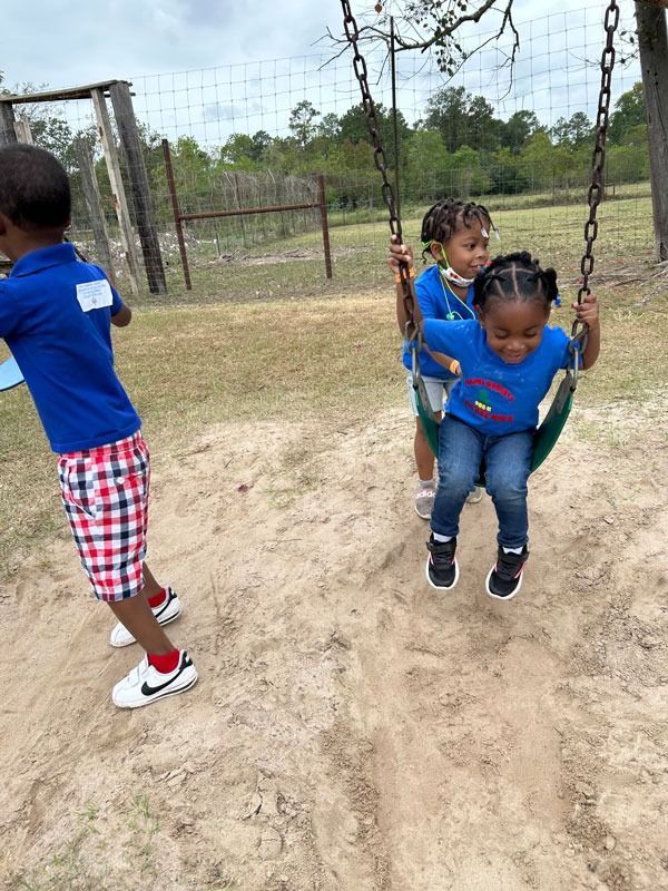 Two children on a swing in a grassy park, with another child standing nearby.
