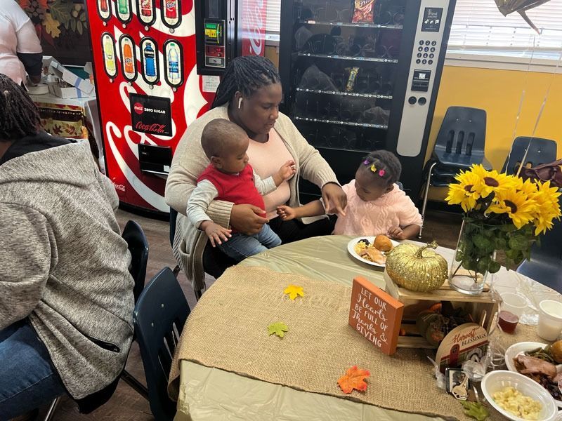 Woman with two children seated at a table in a break room beside a vending machine and flowers