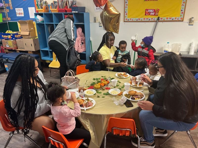 Classroom holiday meal with adults and children seated around a table, sharing food and decorations.
