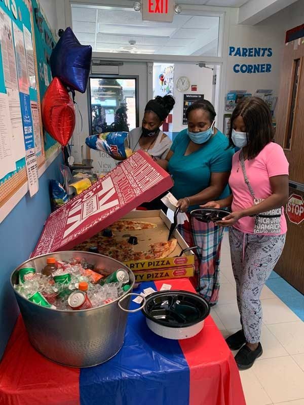 Two masked people behind a snack table in a hallway, with bowls, boxes, and balloons near a Parent's Corner sign.