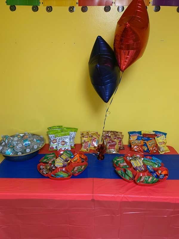 Balloon-decorated table with assorted snacks and treats on red tablecloth against a yellow wall.