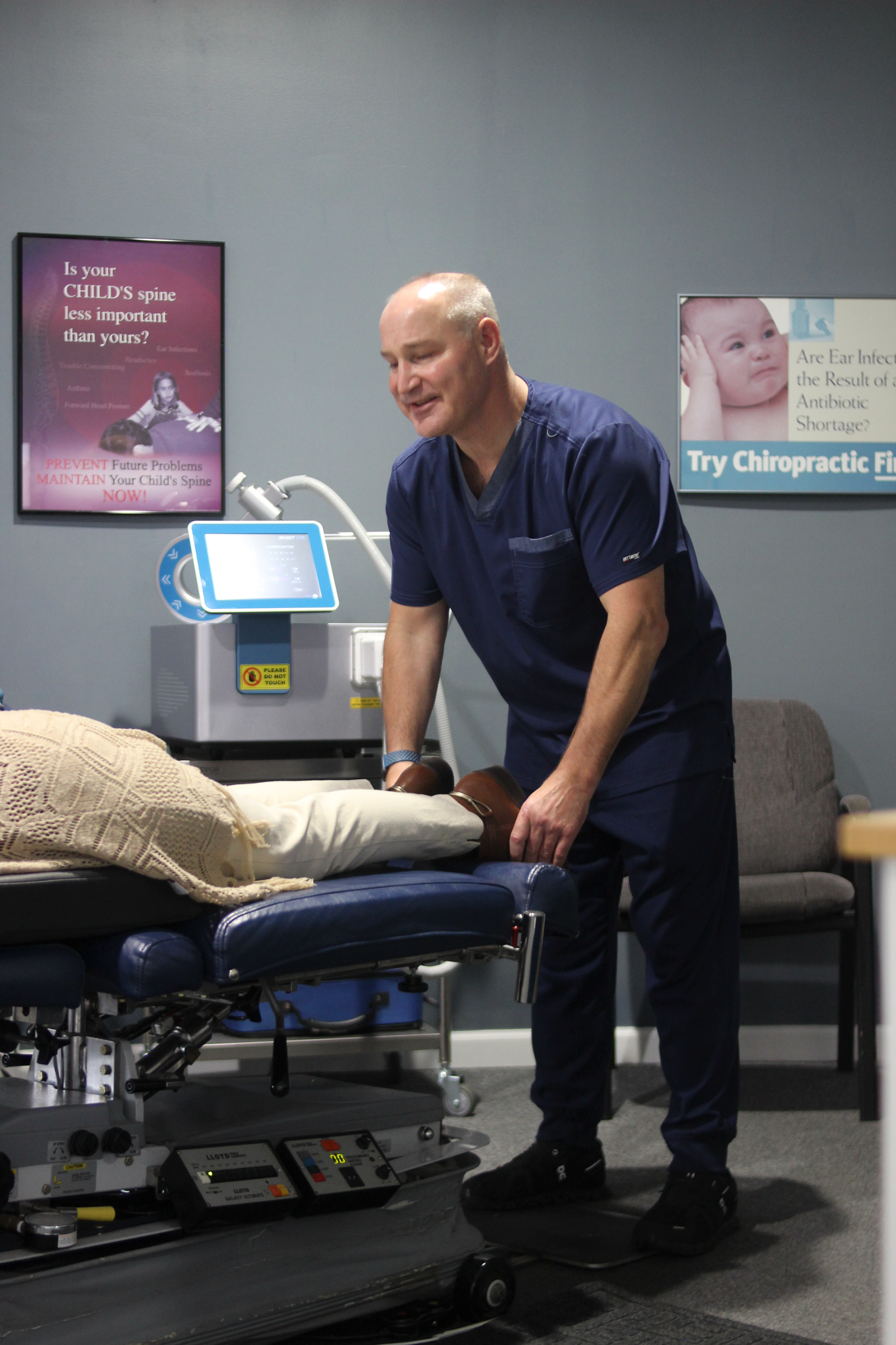 Chiropractor adjusting a patient's leg on a treatment table in an office. The doctor smiles, wearing blue scrubs.