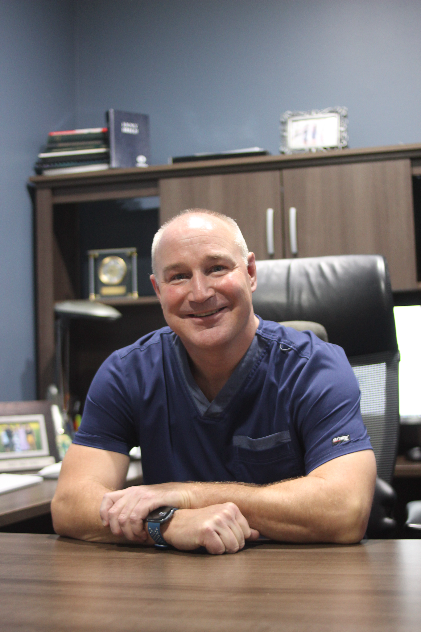 Man in blue scrubs smiling at desk in office.