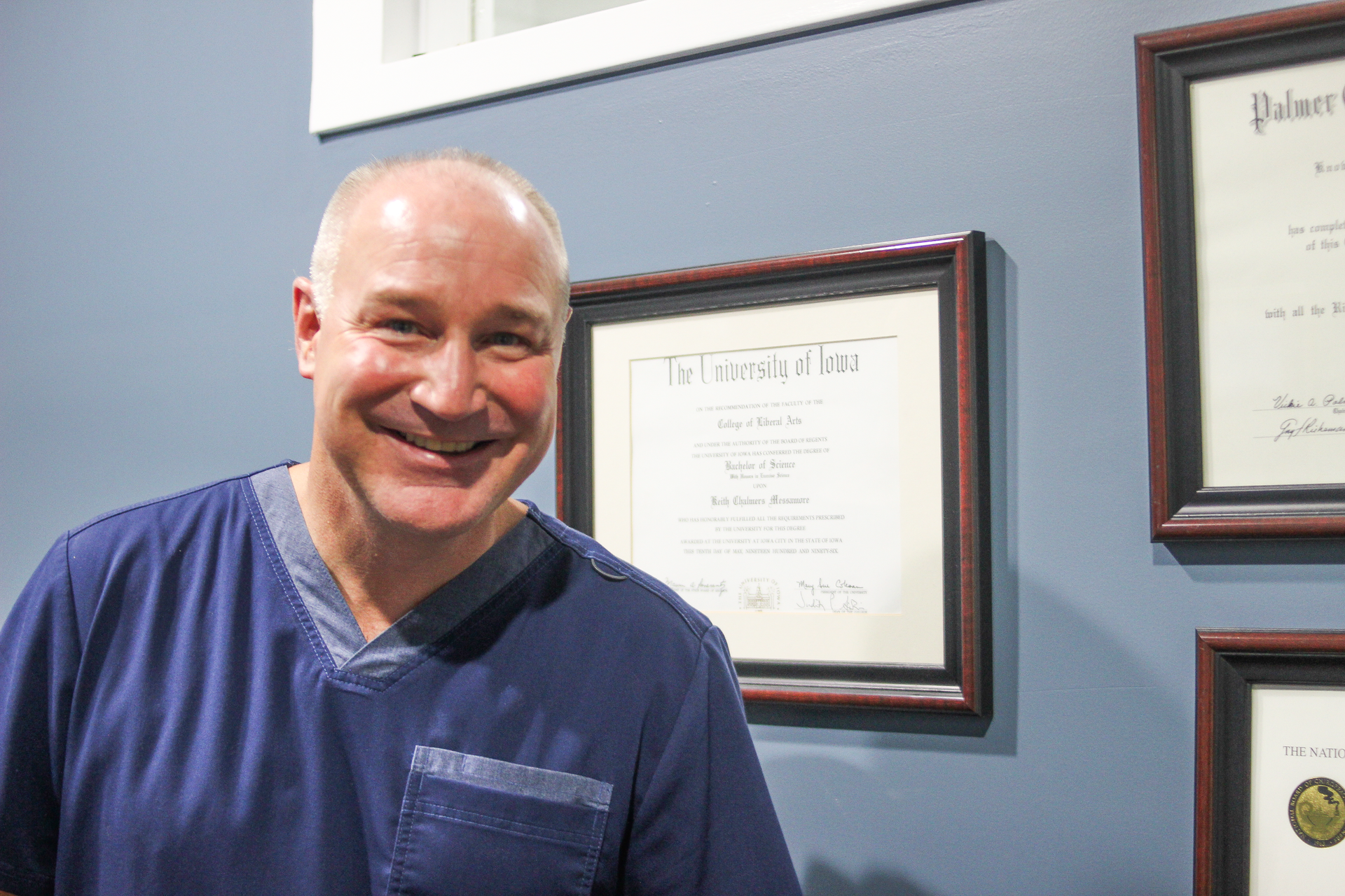 Man in blue scrubs smiles near framed certificates on a blue wall.