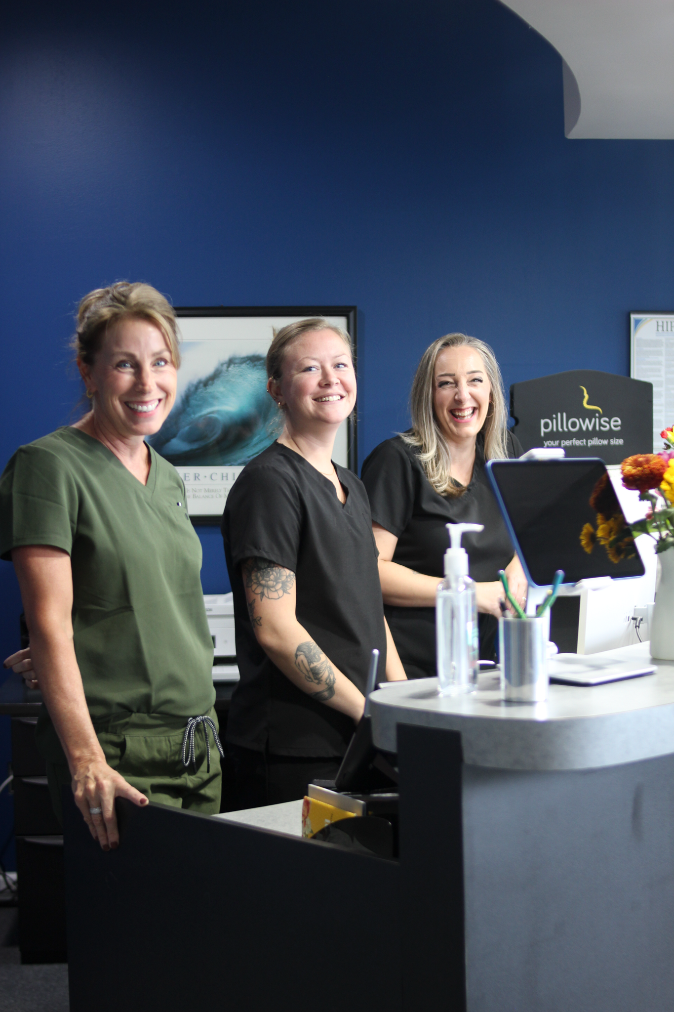 Three people smiling at a reception desk in a dental office. Navy blue wall, logo, and flowers are visible.
