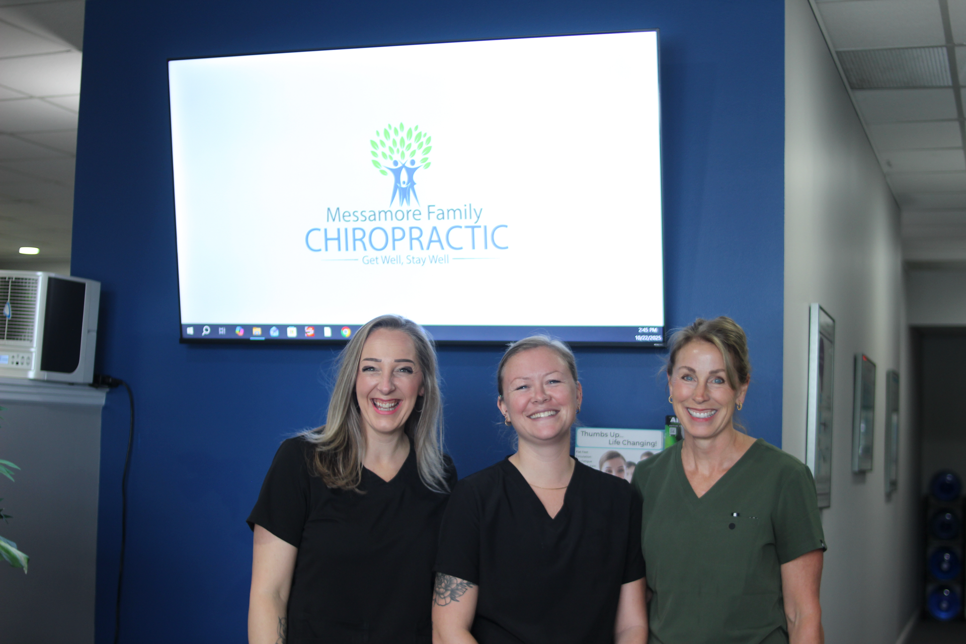 Three women in scrubs stand in a chiropractic office hallway beneath a logo screen.