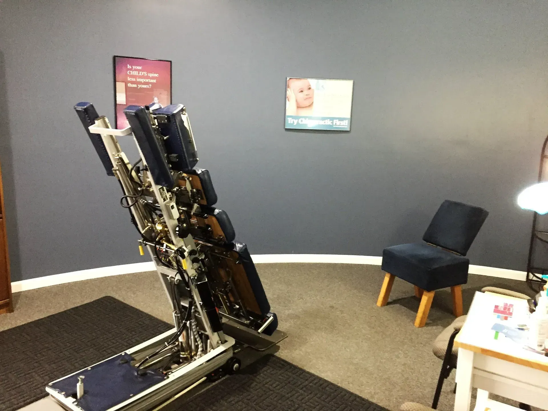 Chiropractic examination room with an adjusting table, chair, and posters on a blue-gray wall.