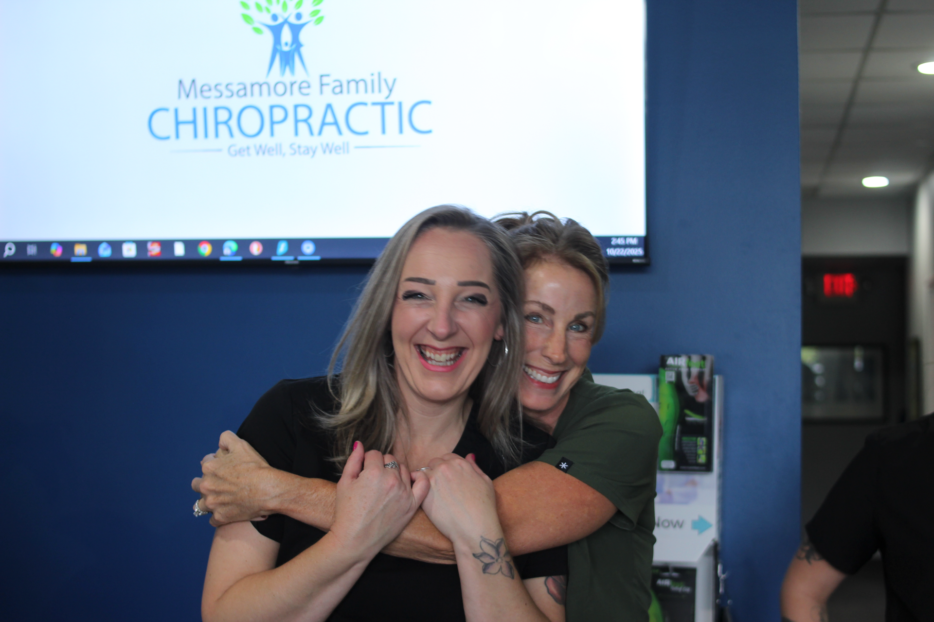 Two women smiling, hugging in front of a chiropractic office sign.