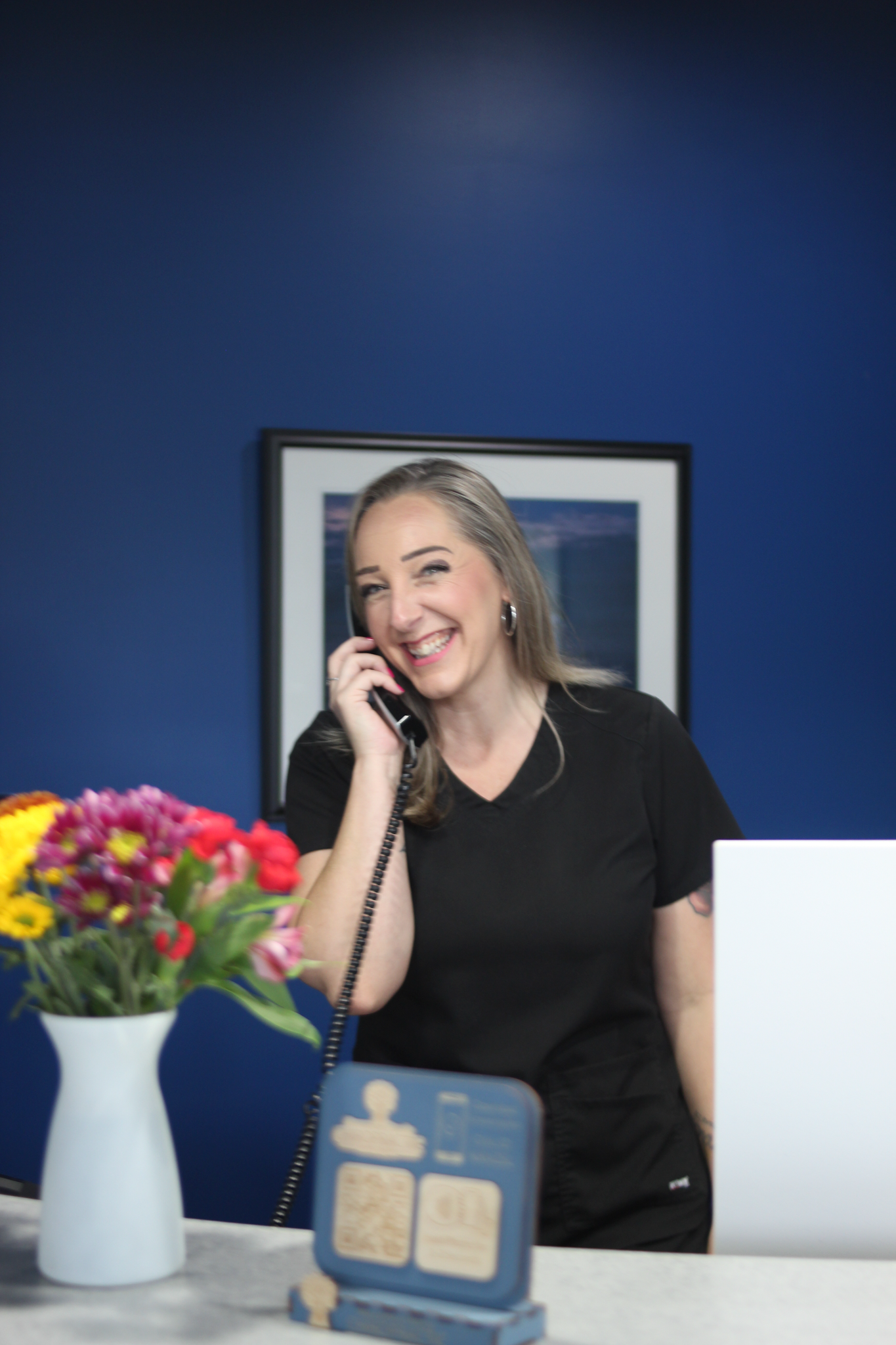 Woman in black shirt smiling while on the phone at a blue desk with flowers and a picture in the background.