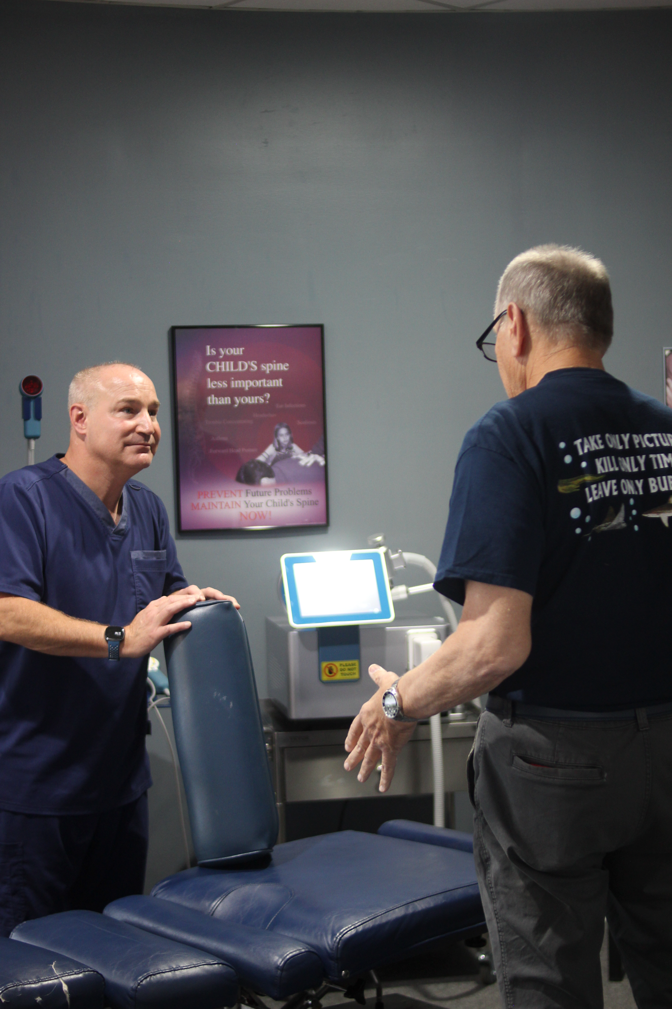 A medical professional in blue scrubs greets a patient in an office with a treatment table.