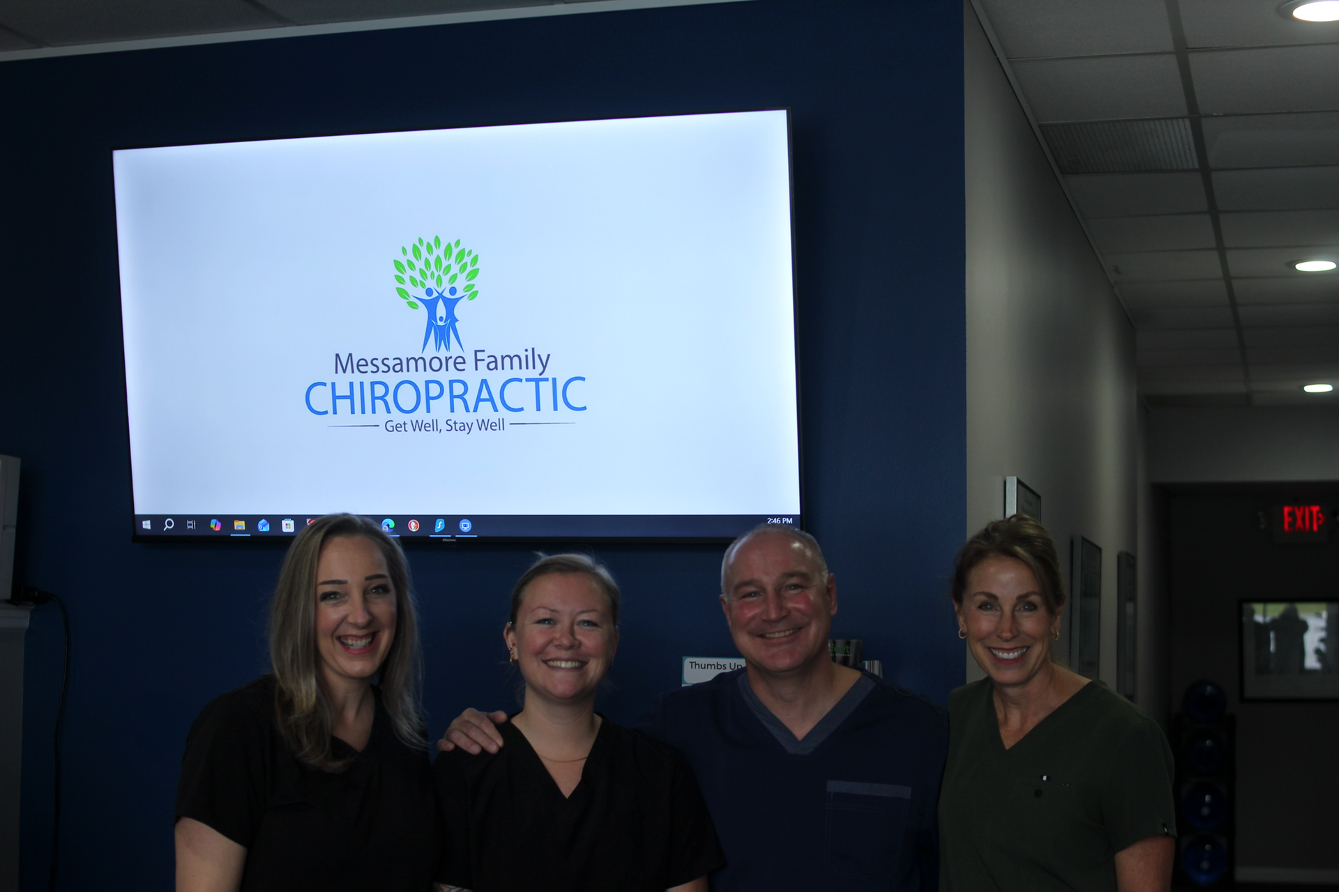 Four people smiling in front of a screen displaying a chiropractic logo; indoors with a blue wall.