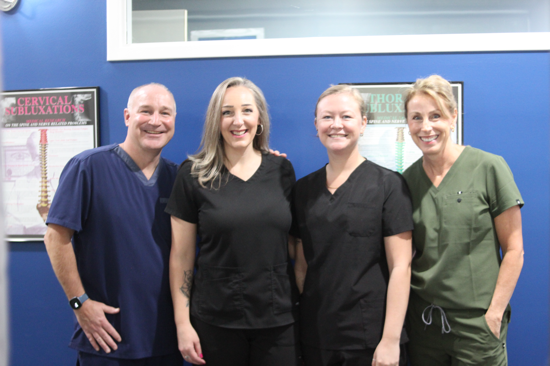 Four people in medical scrubs pose for a photo in a blue-walled office with framed diagrams.
