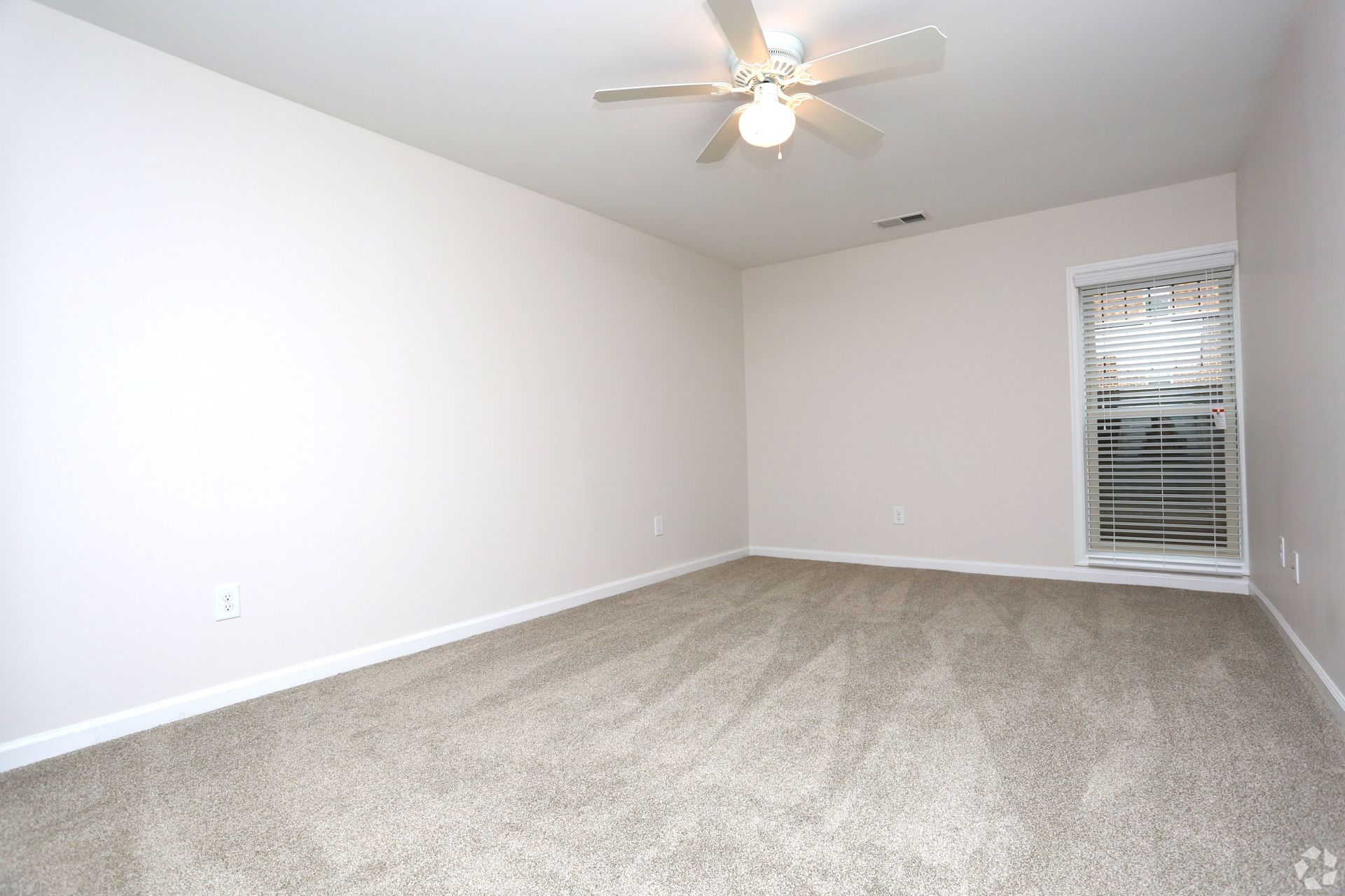 Empty beige carpeted bedroom with white walls, ceiling fan, and a window with blinds