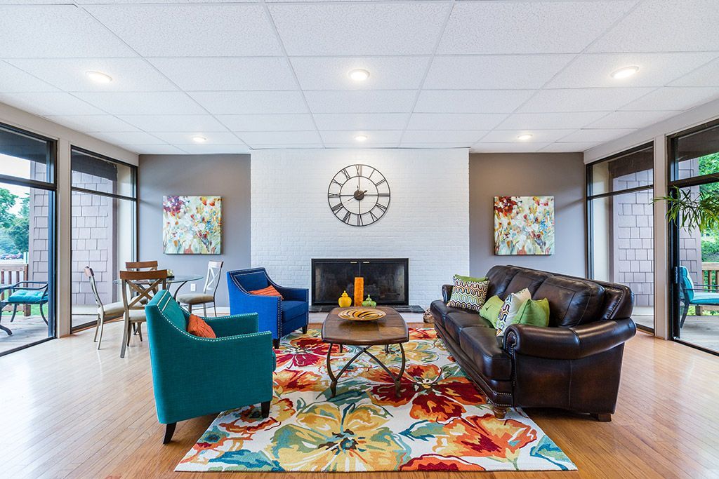 Bright living room with colorful rug, teal and brown chairs, fireplace, wall clock, and large windows
