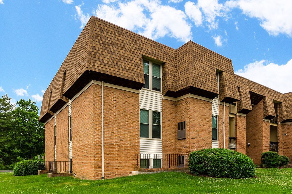 Brick apartment building with steep brown roof, green bushes, and blue sky outdoors