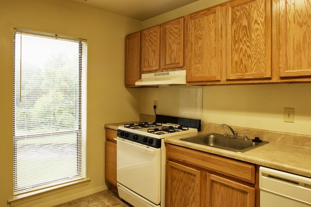 Small kitchen with wood cabinets, beige counters, white stove, sink, and a window with blinds.