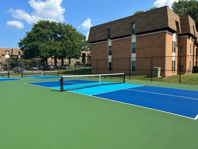 Outdoor tennis court with blue and green surface beside a brick apartment building and trees