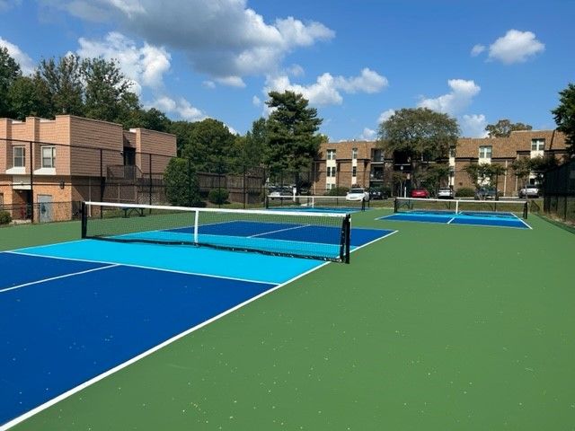 Outdoor tennis courts with blue and green surfaces beside apartment buildings under a partly cloudy sky
