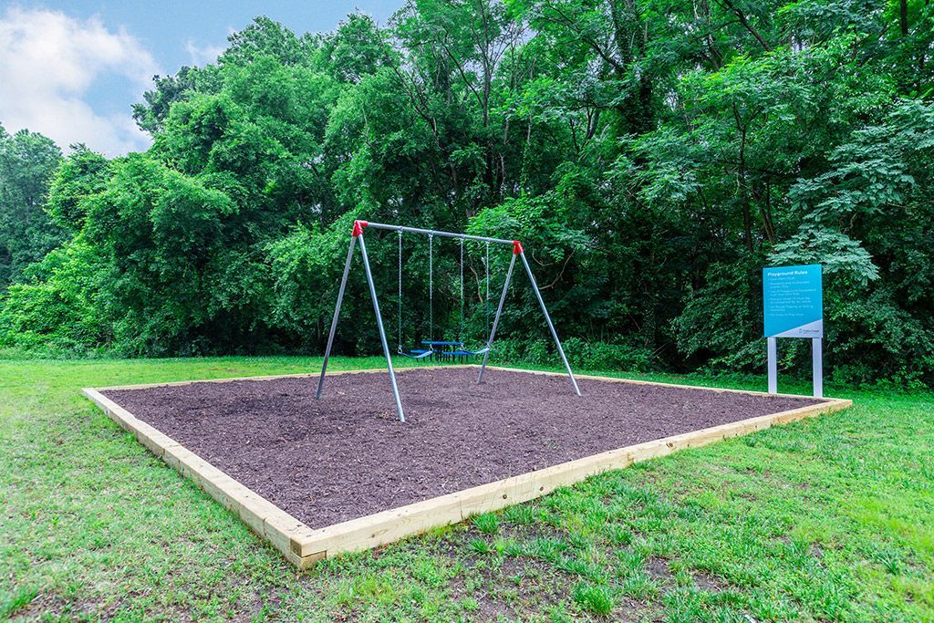 Empty playground swing set beside a dirt court, with green trees and a blue sign.