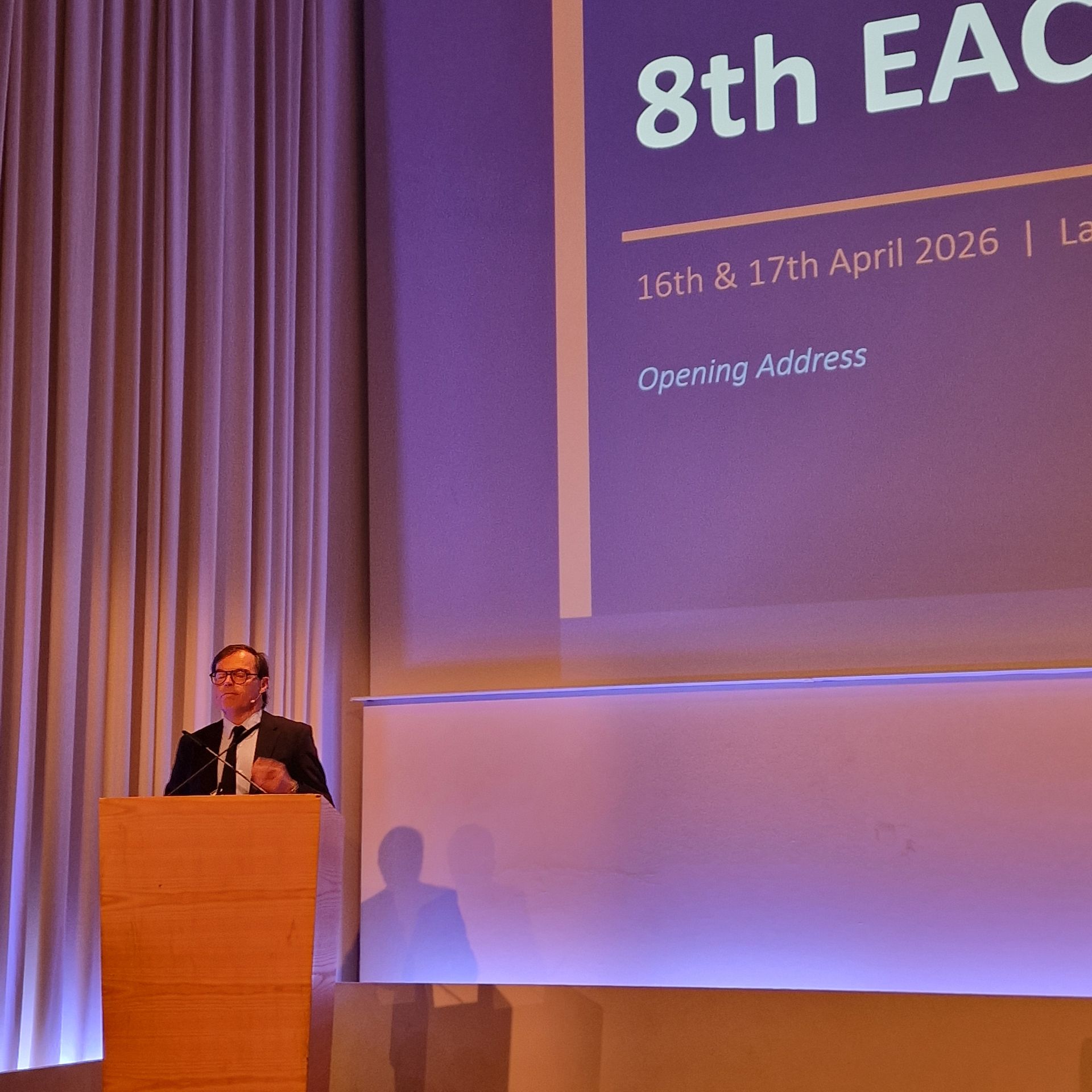 Un orateur prend la parole lors de la 8e conférence de l'EAC, la date de l'événement étant affichée sur un grand écran.