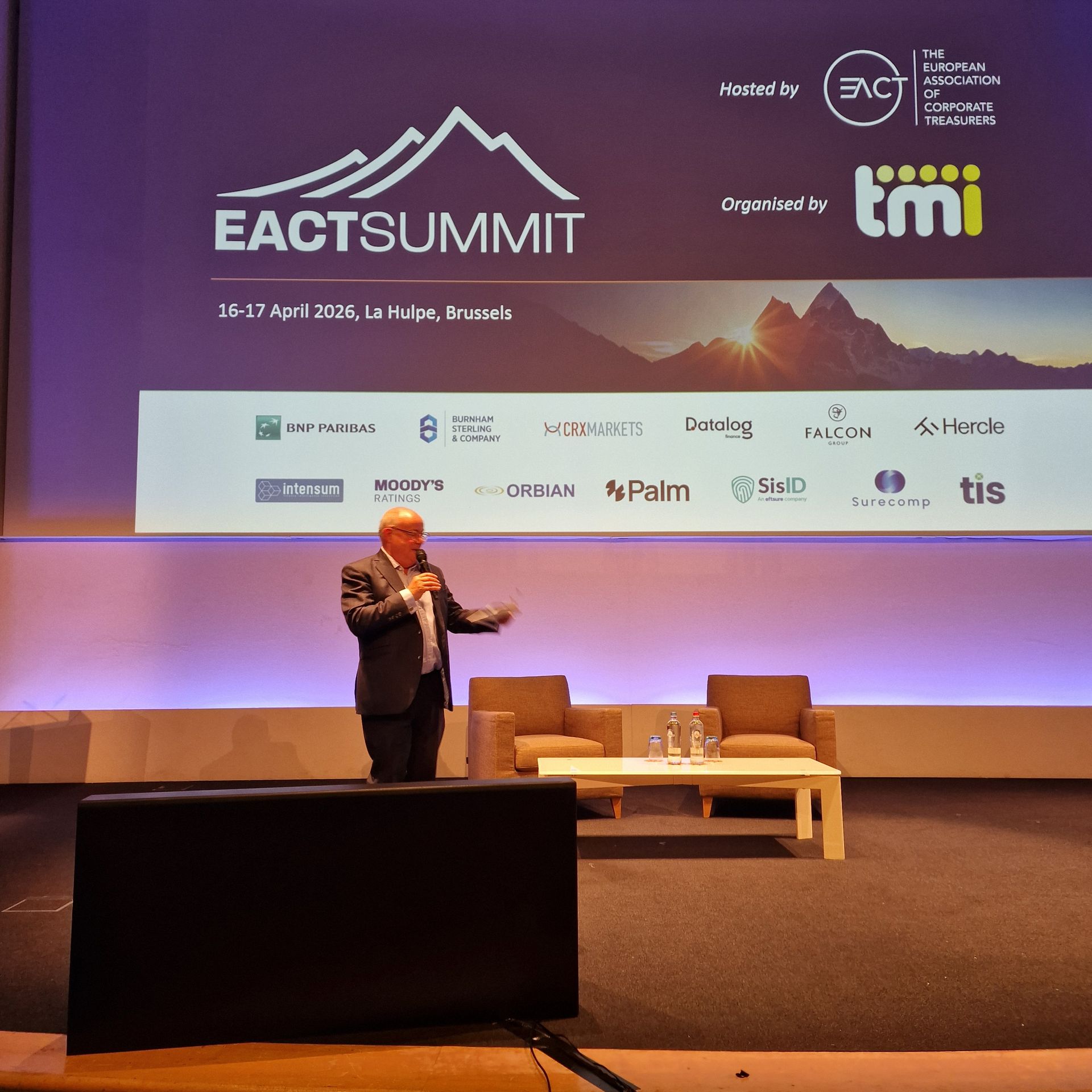 Conférencier sur scène lors de la conférence EACT Summit, avec un grand écran et des sièges derrière lui.