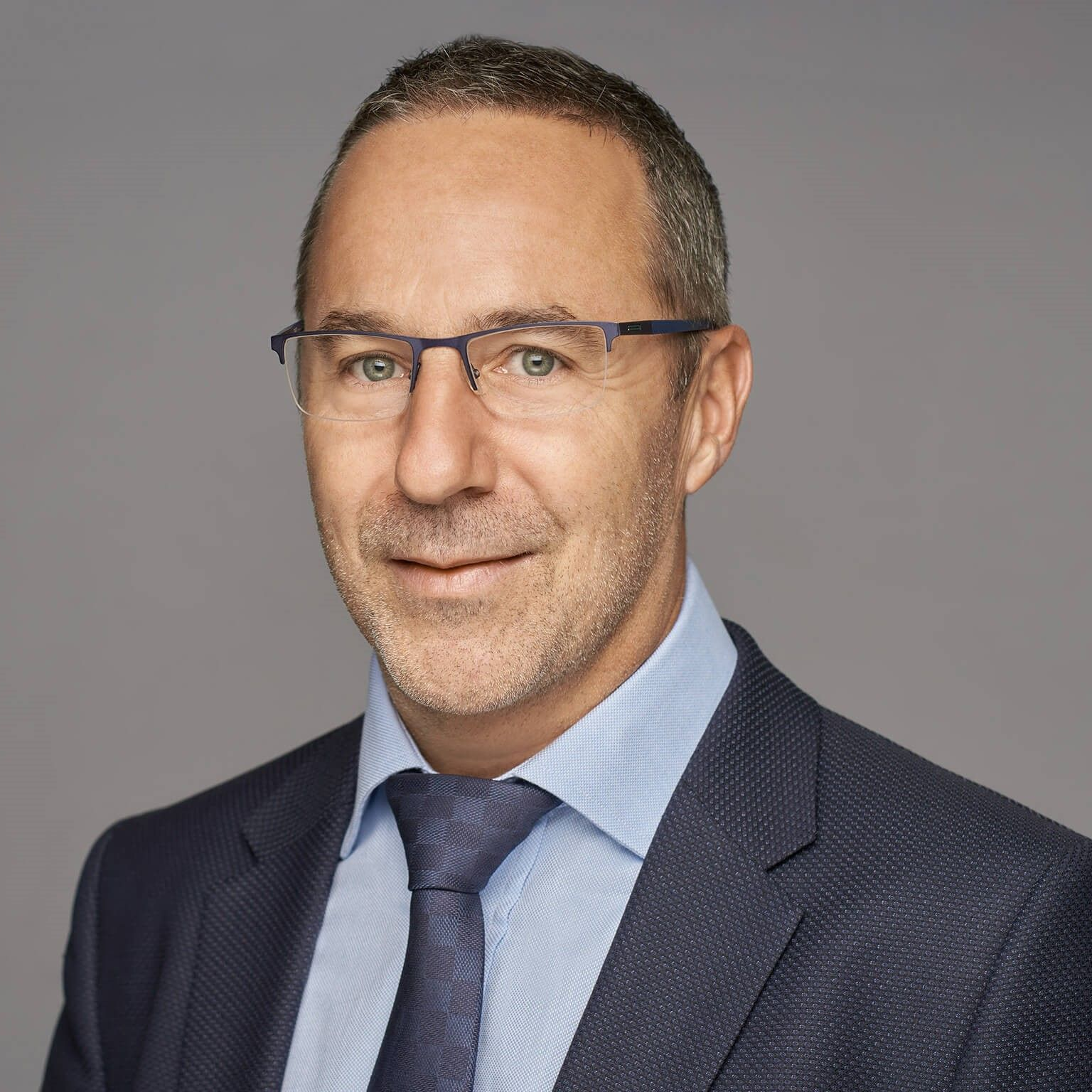 Portrait professionnel d'un homme à lunettes portant un costume sombre, une chemise bleue et une cravate sur fond gris