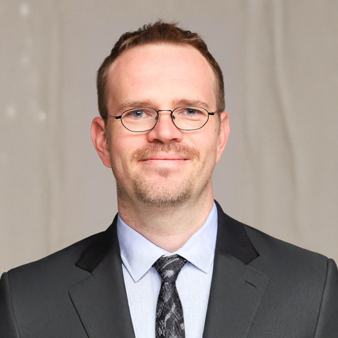 Portrait d'un homme en costume sombre, chemise bleu clair, cravate à motifs et lunettes, souriant sur fond gris.