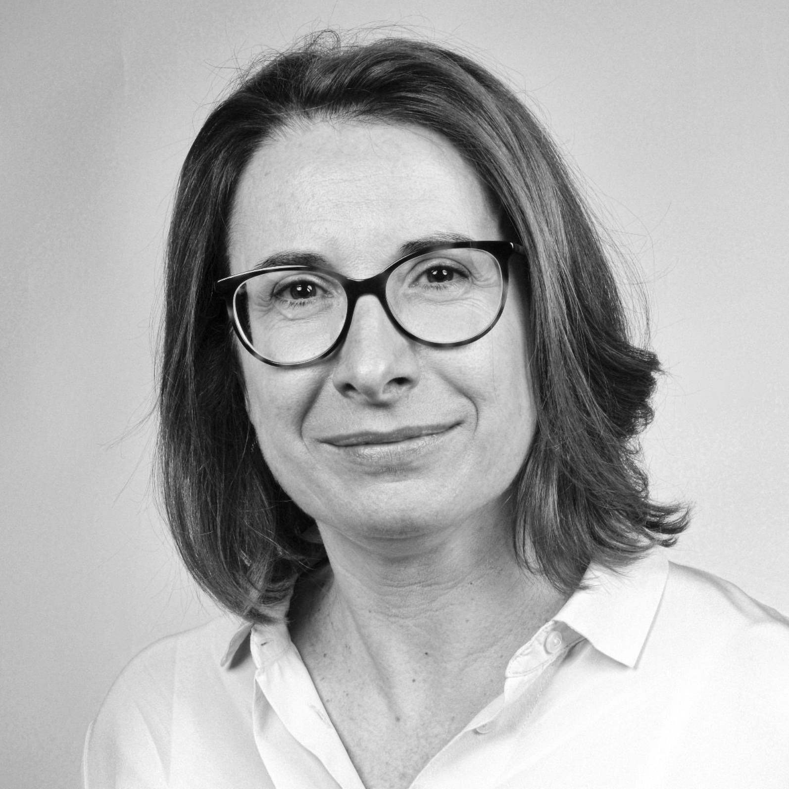 Portrait d'une personne portant des lunettes et une chemise blanche, souriant doucement sur un fond uni