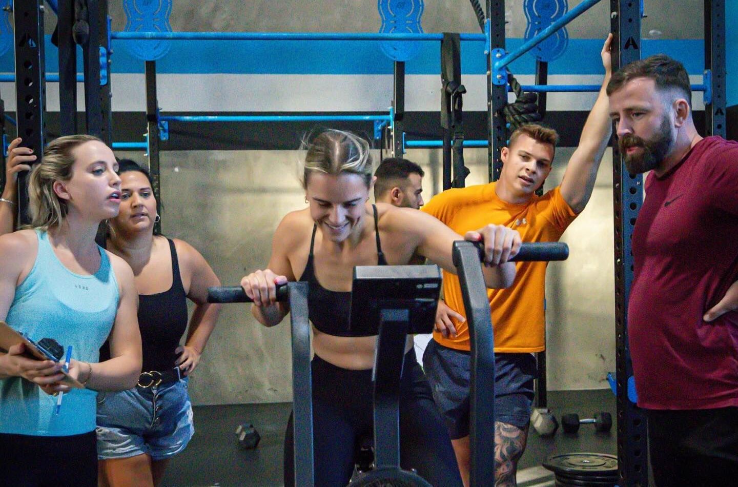 A group of people are standing around a woman riding an exercise bike in Rebuild Health and Fitness in Wynnum West & Capalaba, QLD