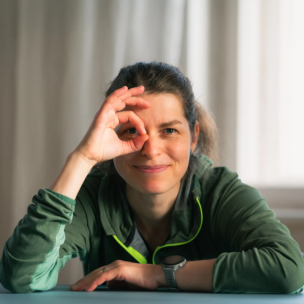 Antje, die mit der Hand vor dem Auge ein „Okay“-Zeichen macht. Sie trägt einen grünen Kapuzenpullover.
