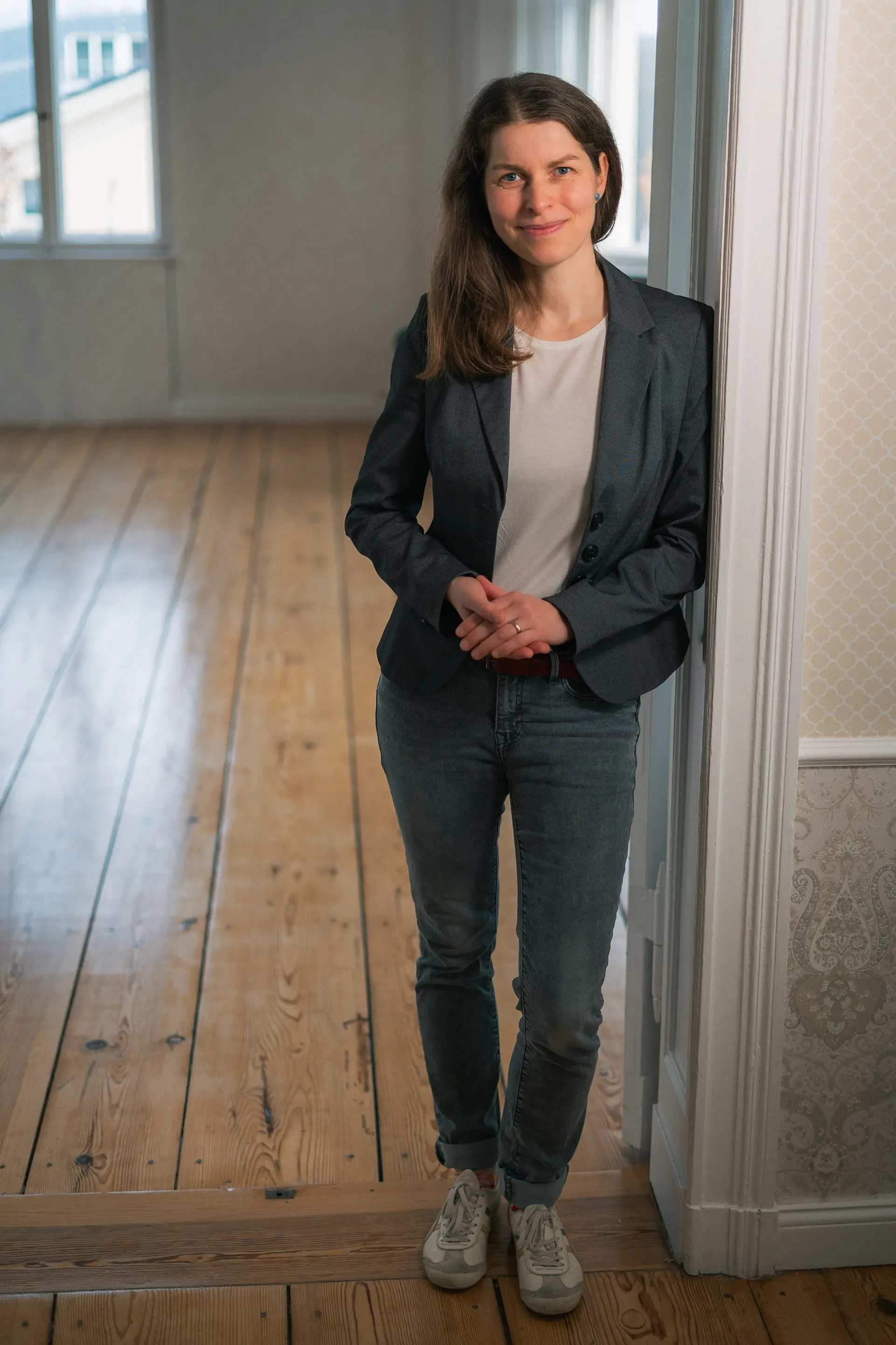 Antje in Blazer und Jeans lehnt in einem hellen Raum mit Holzboden an einem Türrahmen.