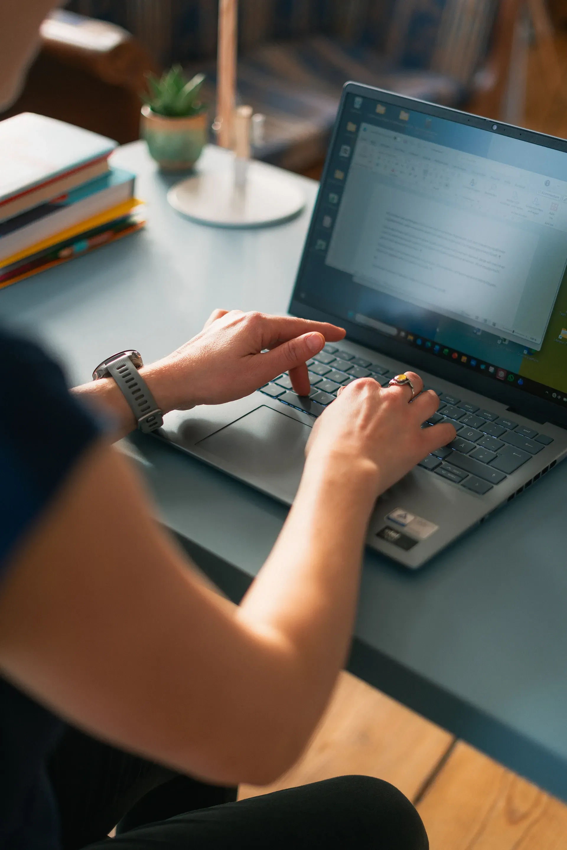 Eine Person tippt an einem Laptop an einem blauen Schreibtisch. In der Nähe befinden sich Bücher und eine Pflanze.