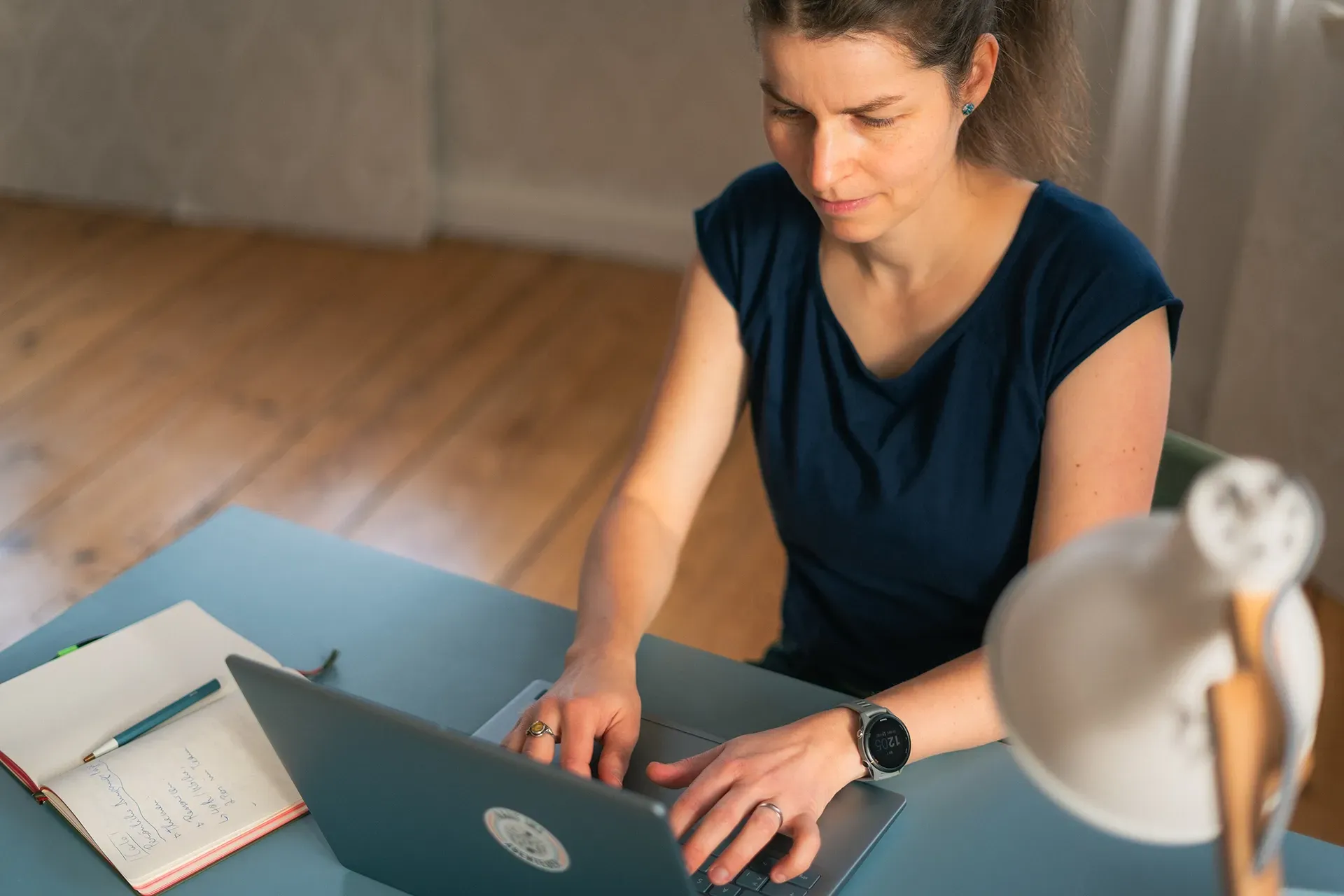 Antje tippt an einem blauen Schreibtisch auf einem Laptop. Notizbuch und Lampe sind zu sehen.