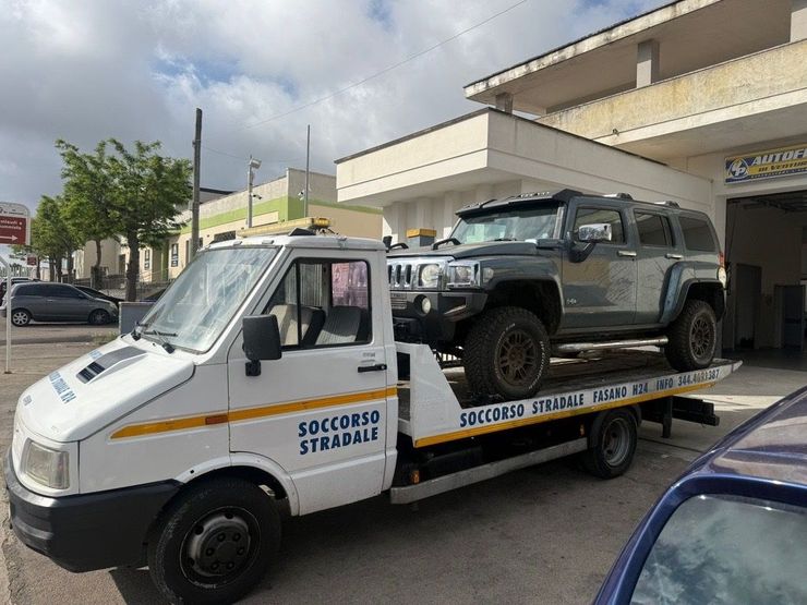 Un carro attrezzi che trasporta un SUV fuoristrada grigio è parcheggiato accanto alla vetrina di un negozio in una via cittadina.