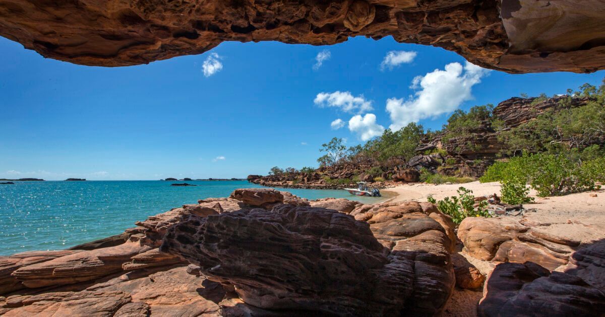 Rocky seaside cove with turquoise water, sandy beach, and a blue sky framed by a cave opening