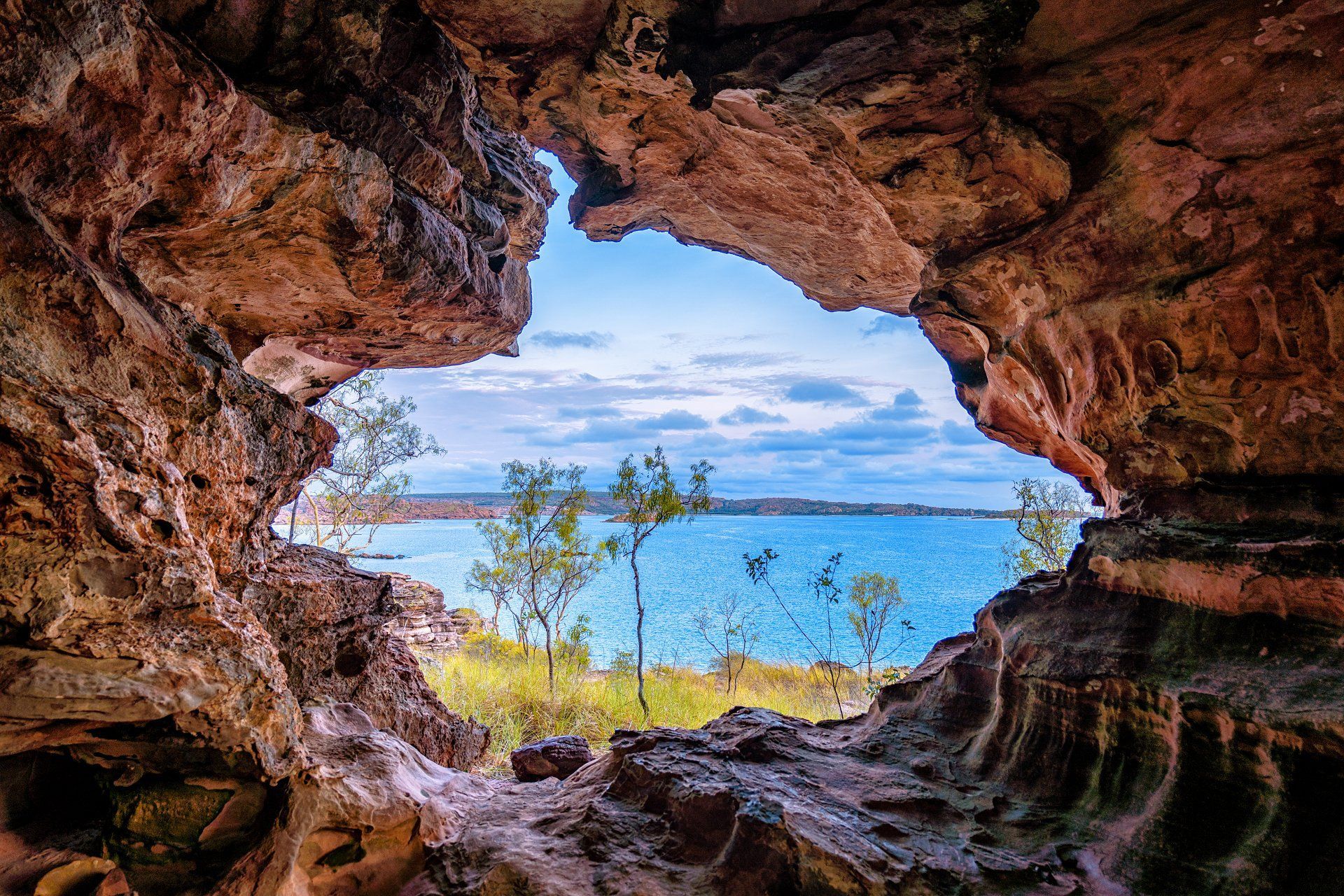 View from a rocky cave opening to a bright blue lake and sky, with trees in the foreground