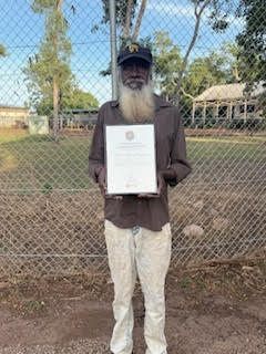 Man with long beard holds a framed document, standing in front of a chain-link fence, outdoors.