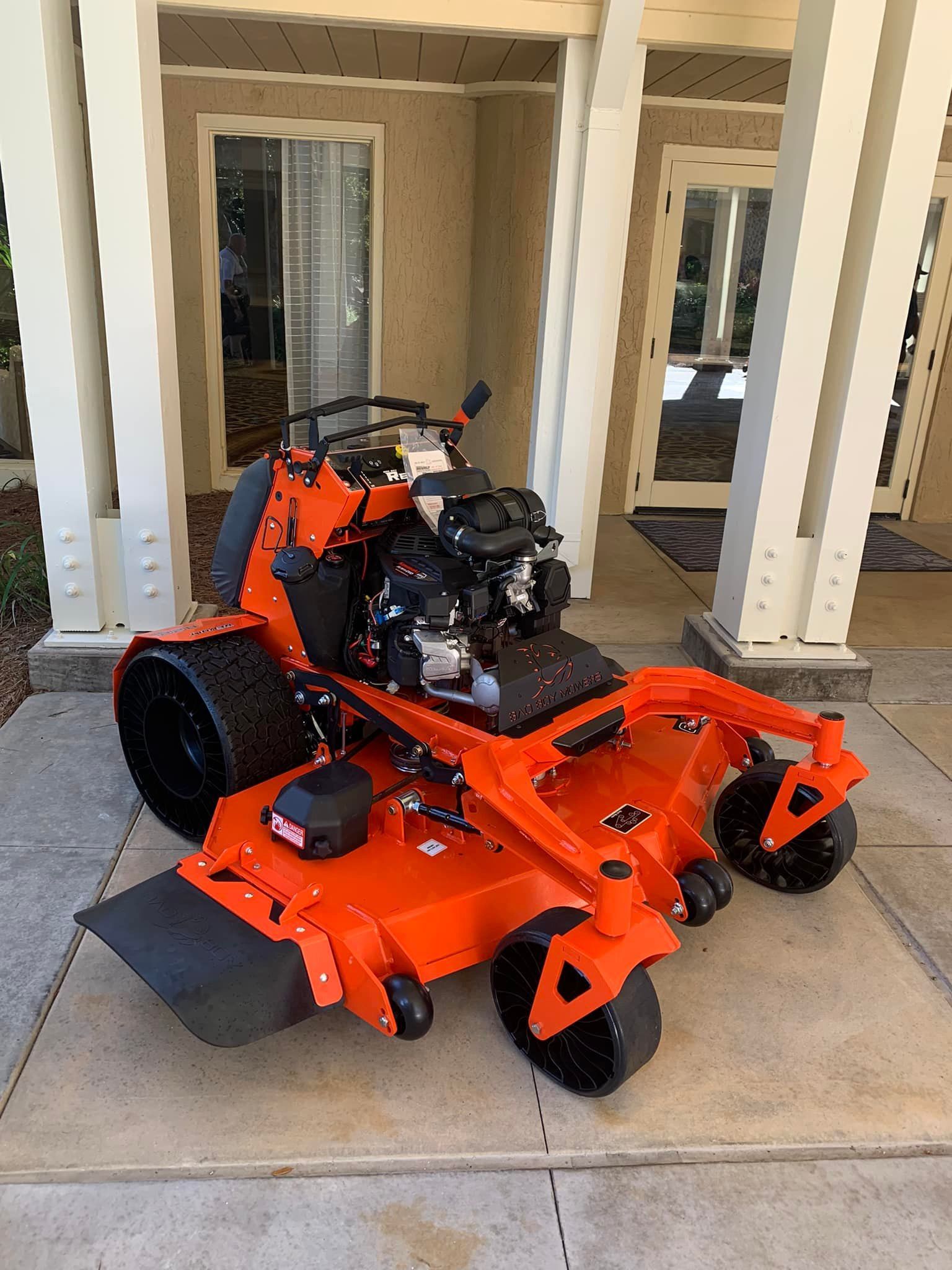 An orange lawn mower is parked on the sidewalk in front of a house.