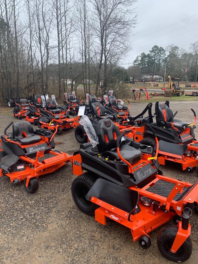 A bunch of orange and black lawn mowers are parked in a lot.