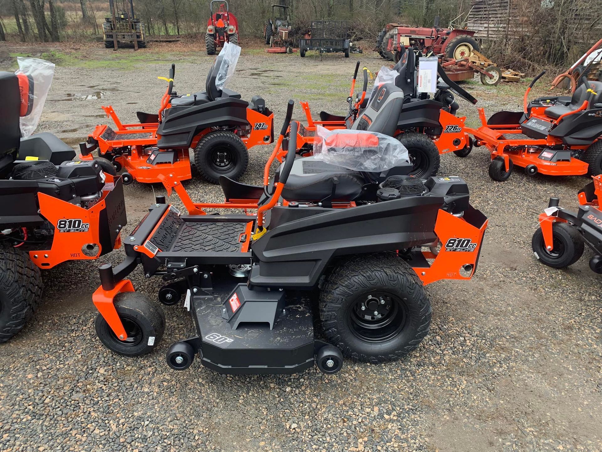 A row of orange and black lawn mowers are parked in a gravel lot.