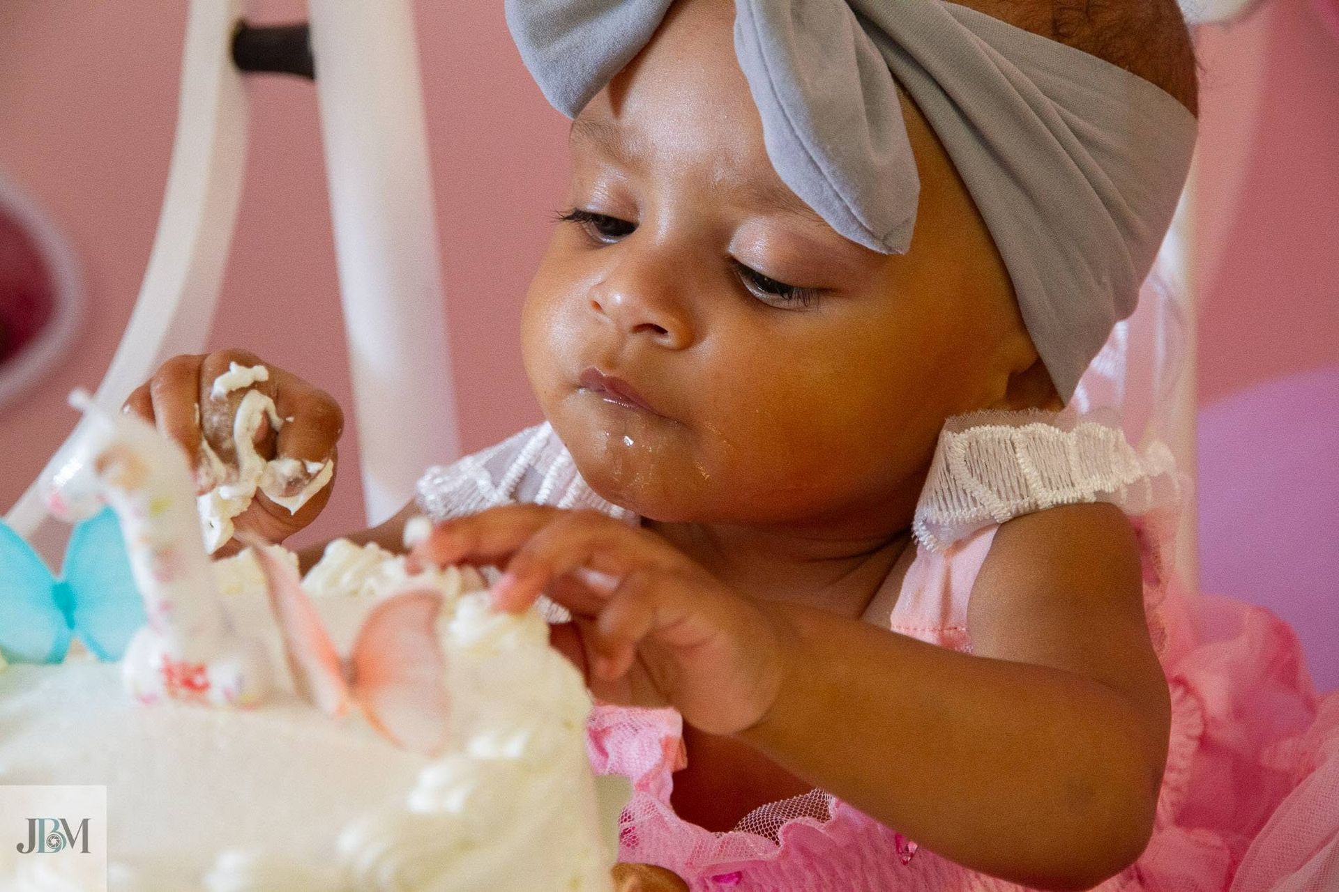 Toddler in a gray headwrap reaching for cake at a pink birthday celebration