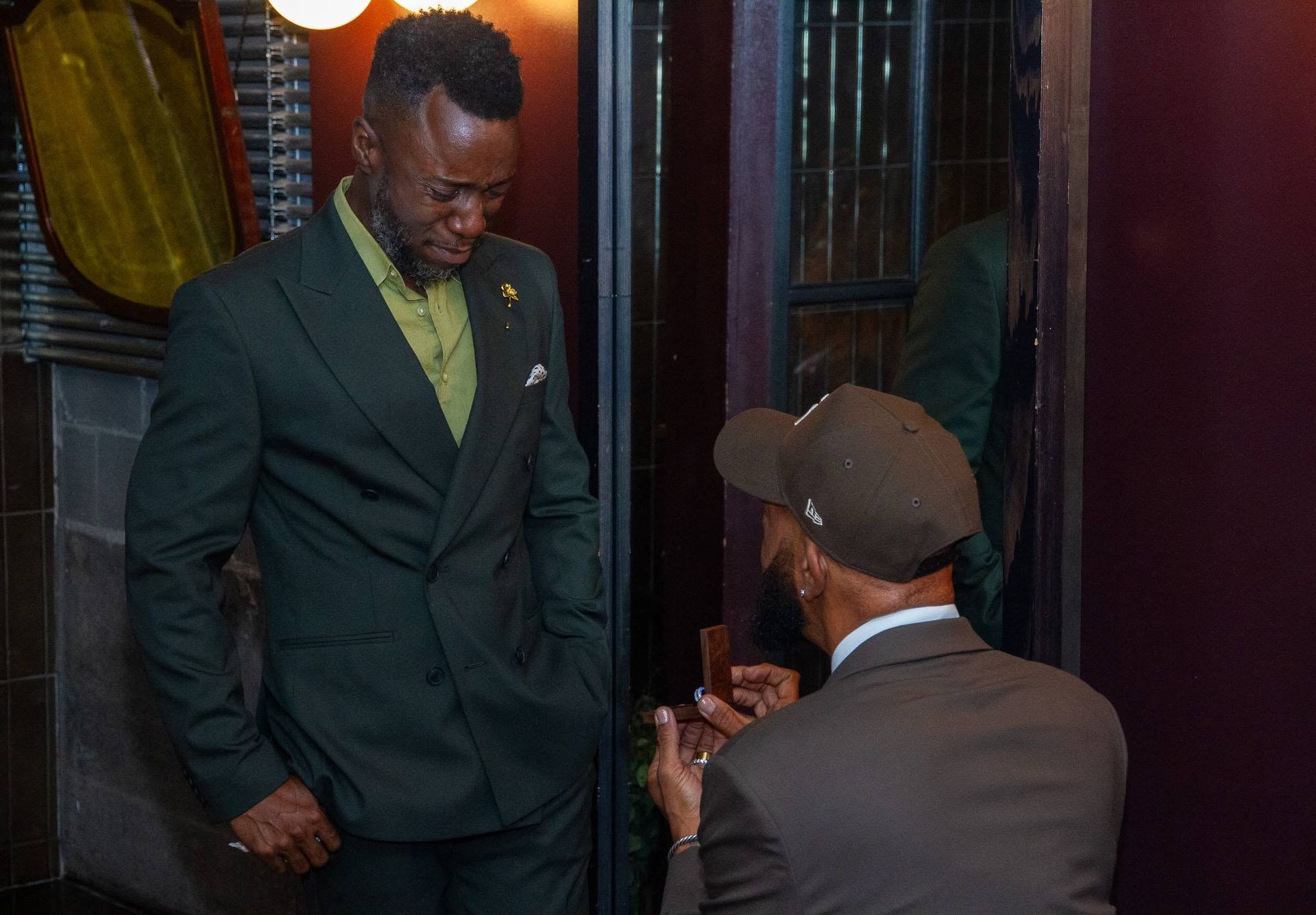 Two men in formal wear conversing in a doorway, one standing and one seated, in a dim indoor setting.