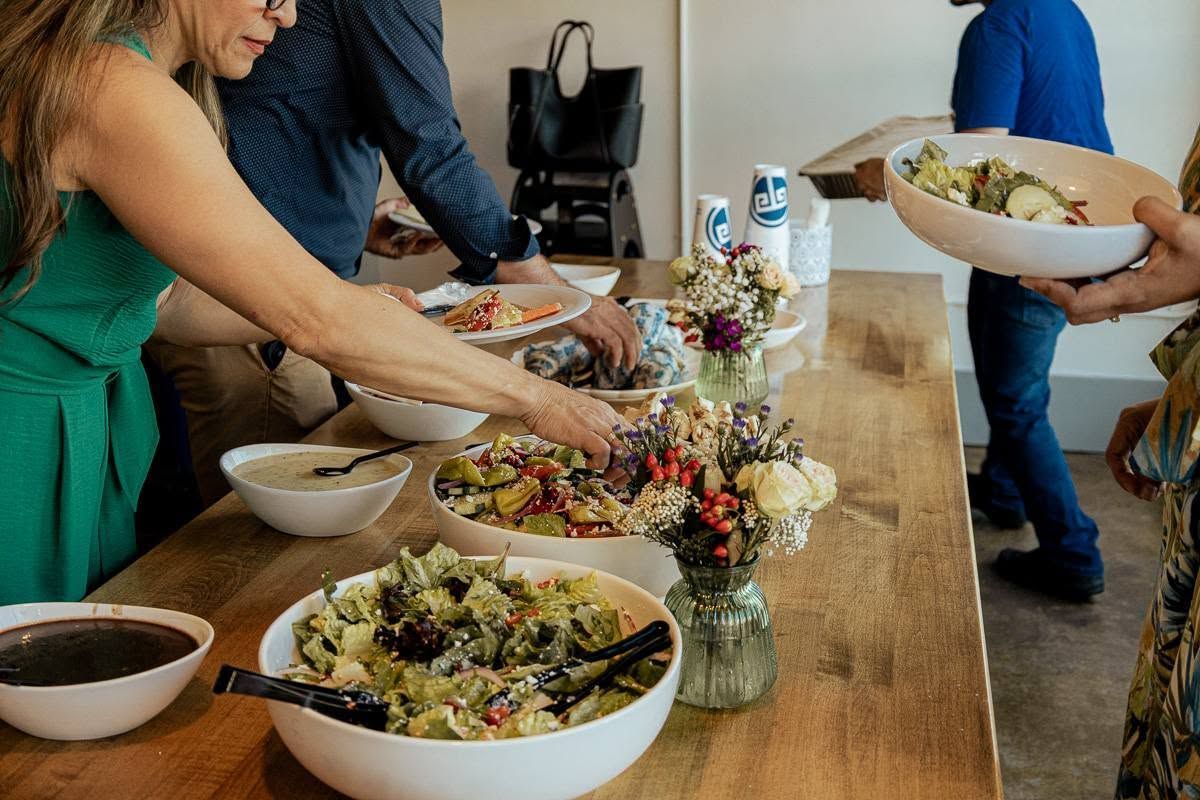 People serving food buffet-style on a wooden table with salad bowls and flowers