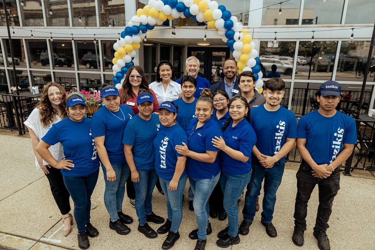 Group of people in blue shirts posing under a blue, white, and gold balloon arch outside a building