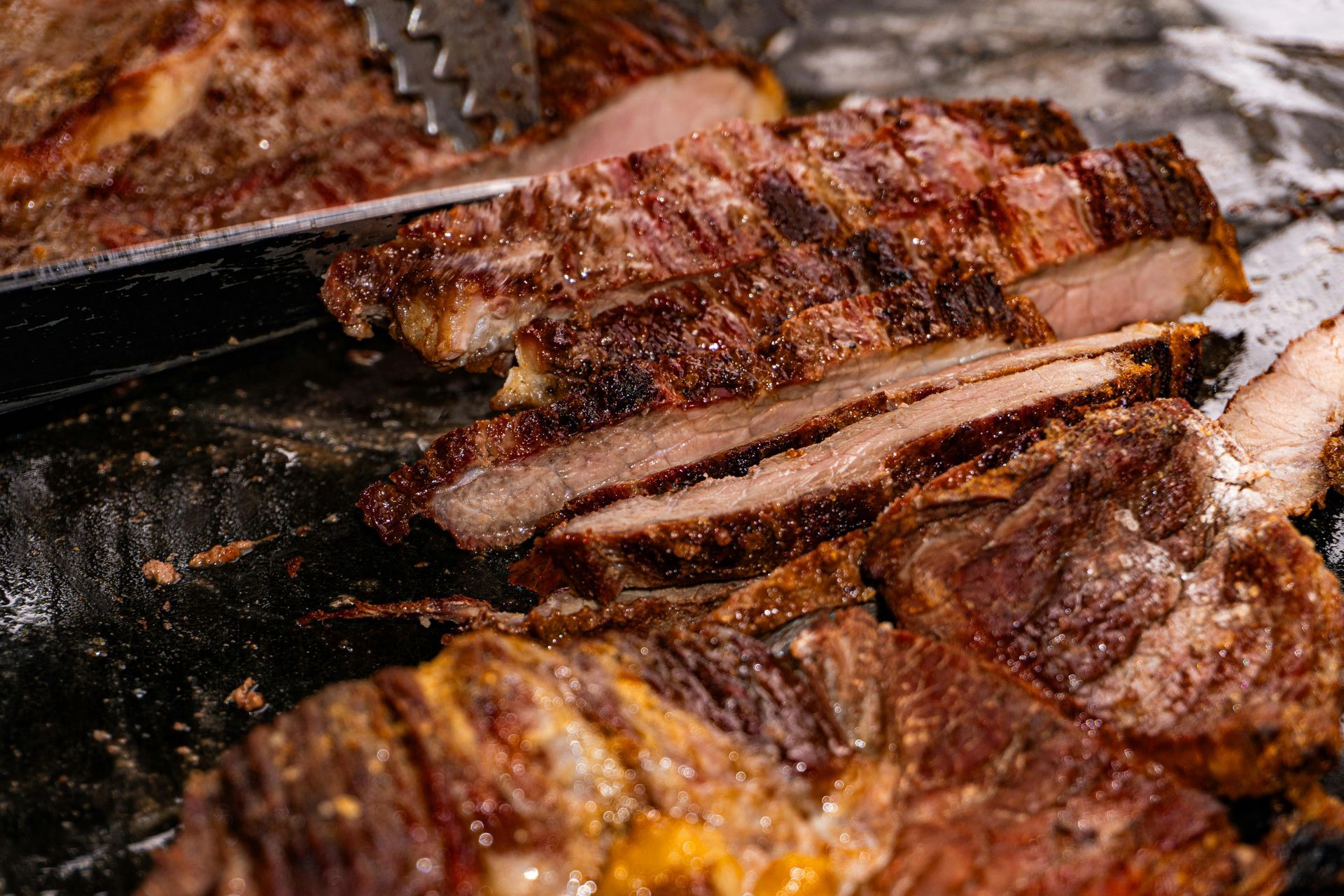 Sliced grilled steak, close-up. Charred exterior, pink interior. Shown with tongs.