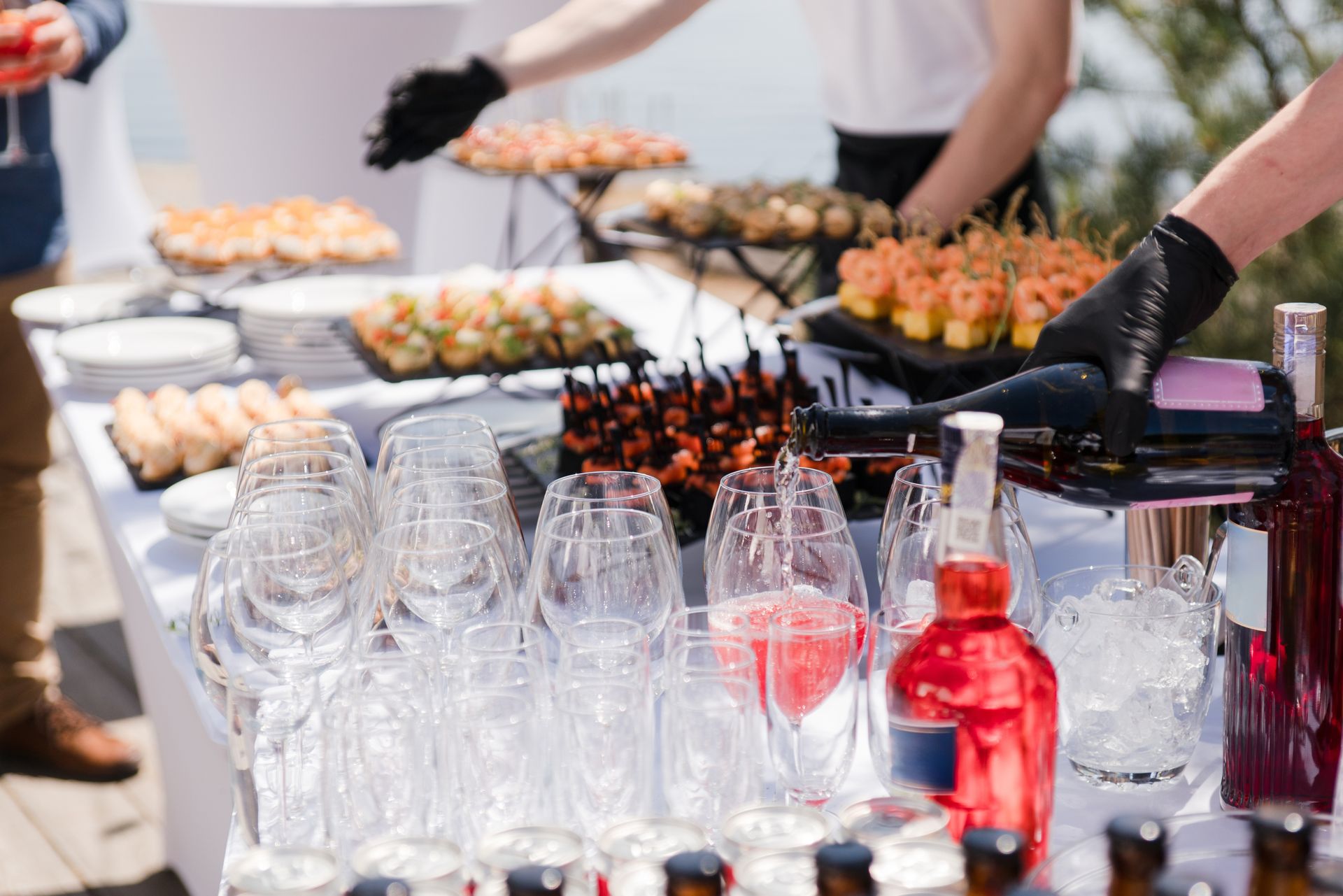 Outdoor buffet table with stacked glasses and red drinks, while servers arrange appetizers in the background